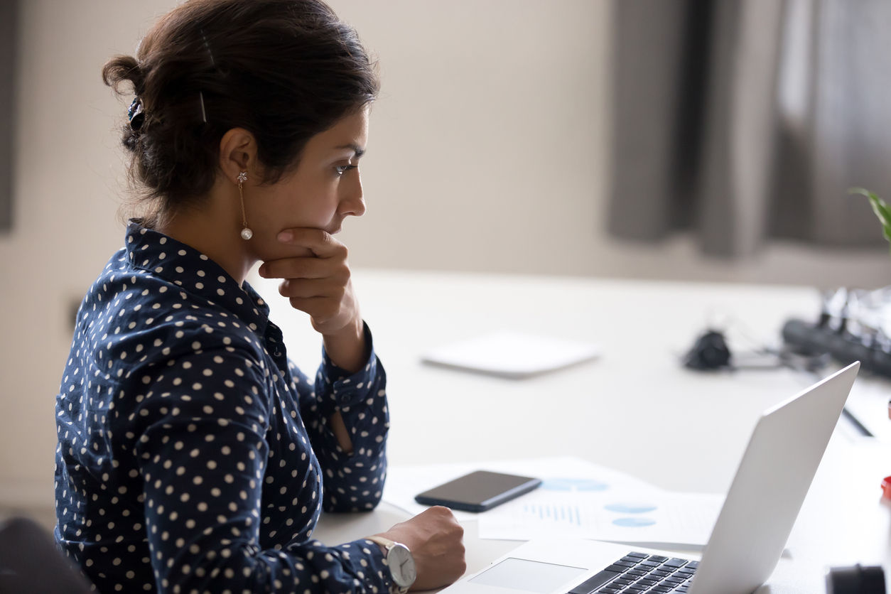 Indian girl sitting at desk near computer cogitating thinking making important decision at workplace. Concentrated serious office worker millennial woman analysing results feels doubts and feel unsure