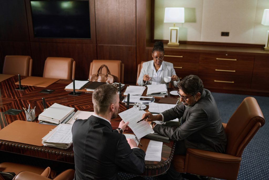 Mature boss in formalwear pointing at financial or juridical paper document while explaining its points to male colleague